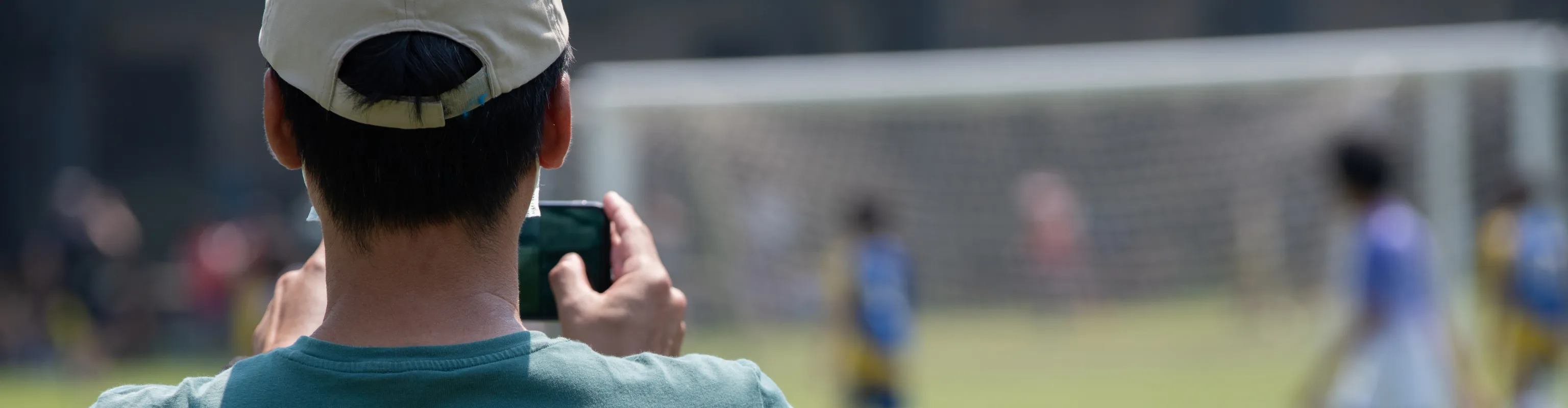 Man in white hat and green tshirt, facing away from camera, taking a photo of a kids soccer game