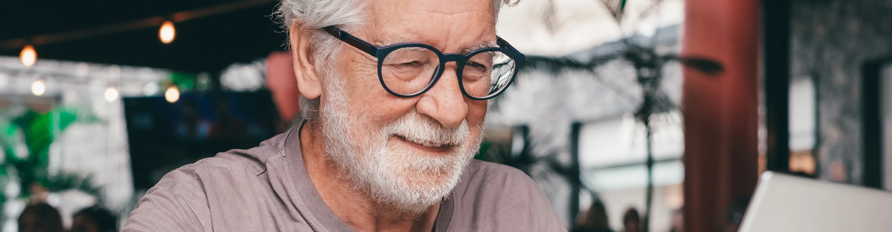 A man with silver hair and glasses smiles and works away on his laptop and smartphone