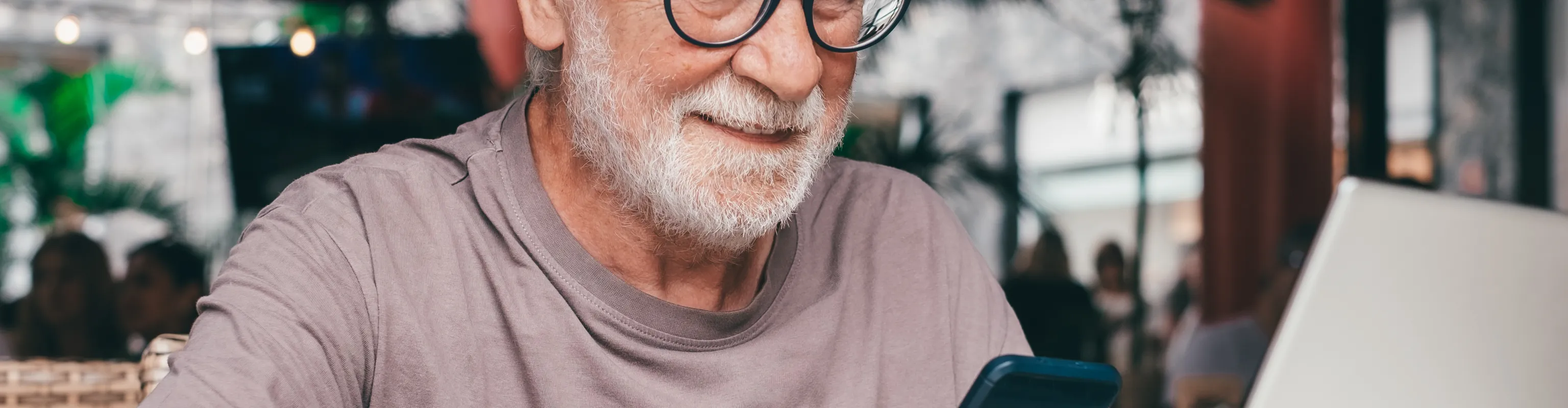 A man with silver hair and glasses smiles and works away on his laptop and smartphone
