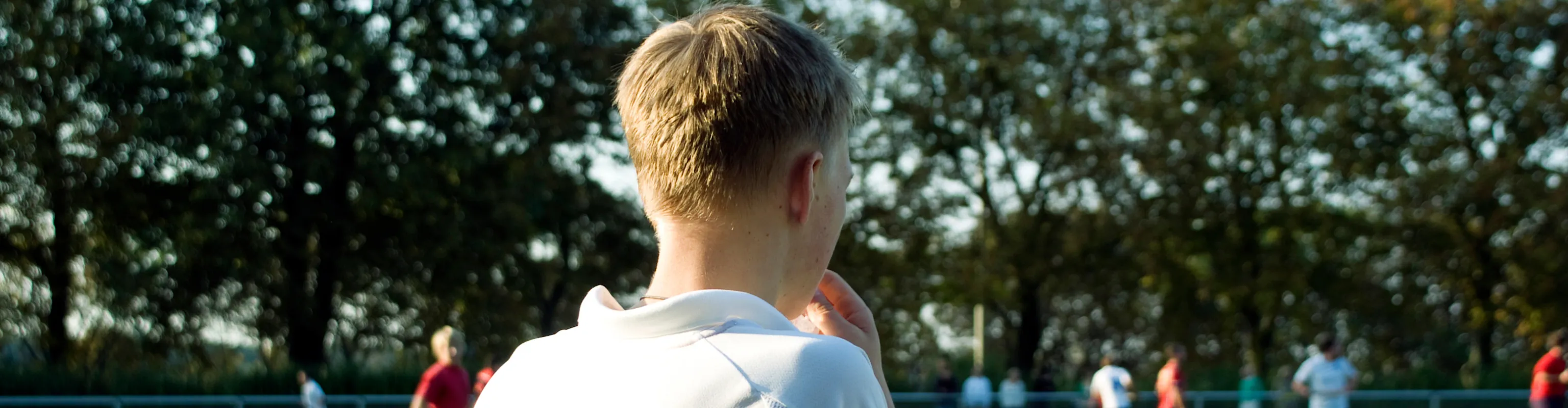 A teenage boy stands by the sidelines of a field hockey game with his back to the camera. All the action is far in the background.