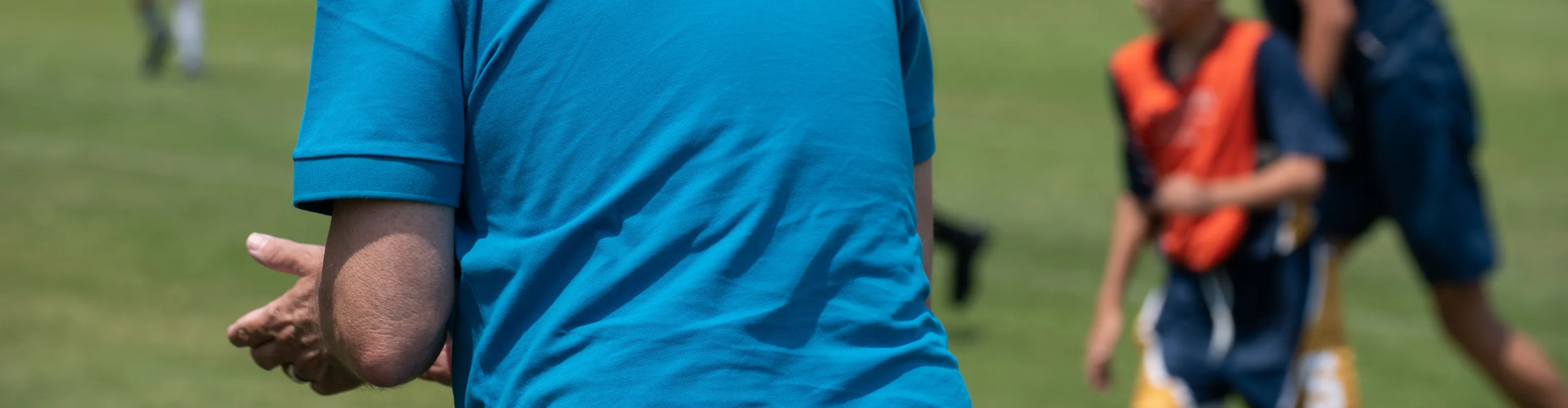 A parent on the sidelines claps for their child as they play football. The parent is wearing a blue polo shirt and the kids are blurred by the depth of field.