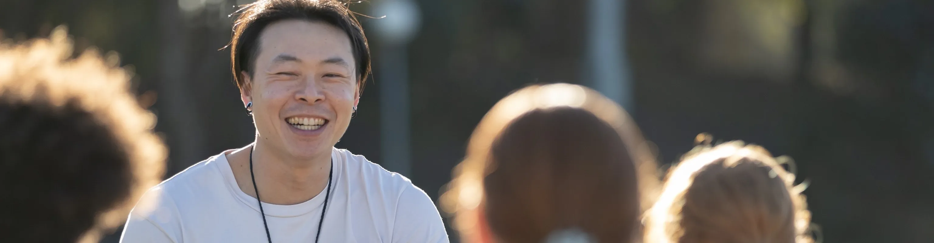 A male coach of East Asian appearance in a white tshirt holds an iPad and smiles while talking with female athletes who are listening attentively