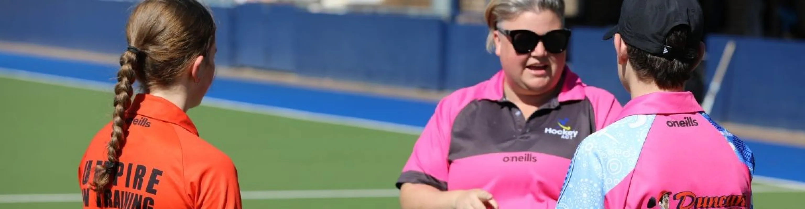 A match official talks to to a junior official as a girl wearing an 'umpire in training' polo shirt