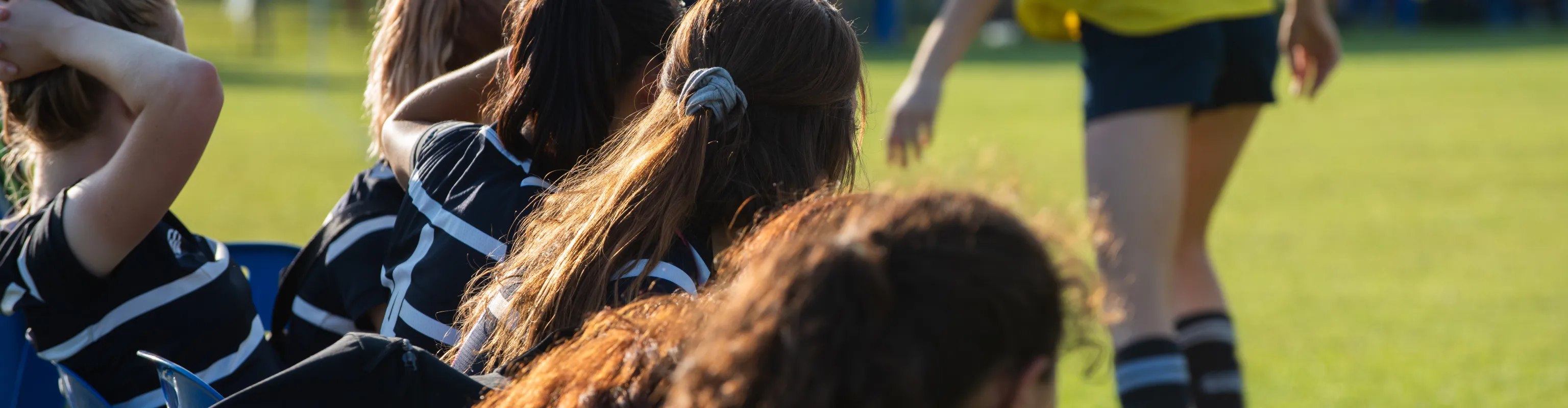 Girls on a rugby team sit on the bench while observing the field.