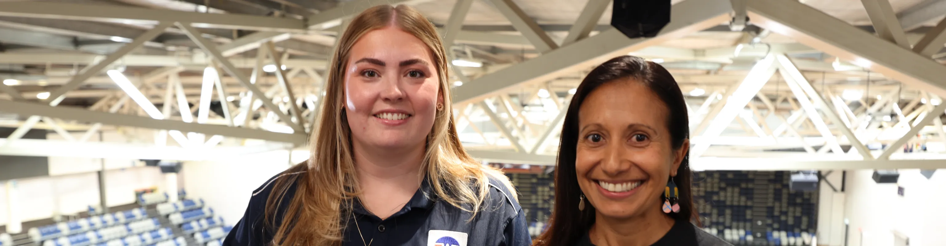 Two women smile at the camera inside an indoor stadium.