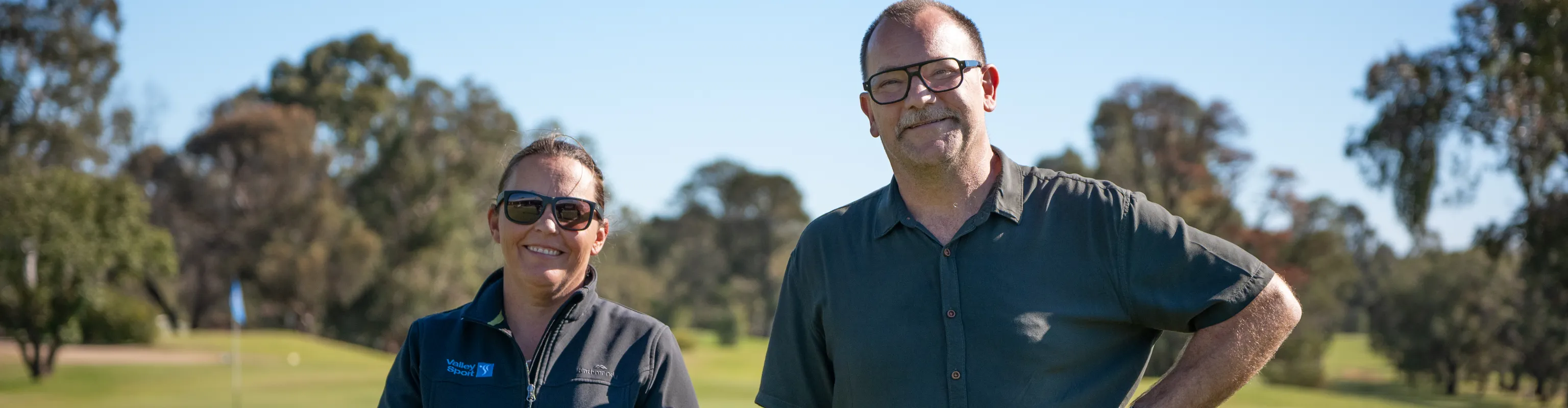 One women and one man with fair skin stand smiling on a golf course in the sun.