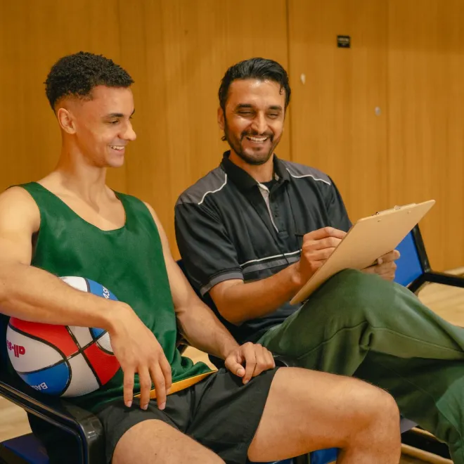 A coach and player sit on the sidelines of a basketball court smiling