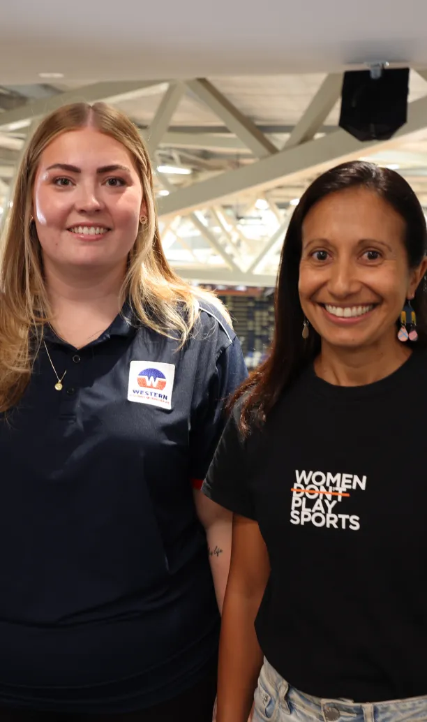 Two women smile at the camera inside an indoor stadium.