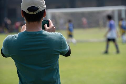 Man in white hat and green tshirt, facing away from camera, taking a photo of a kids soccer game