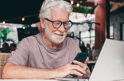 A man with silver hair and glasses smiles and works away on his laptop and smartphone