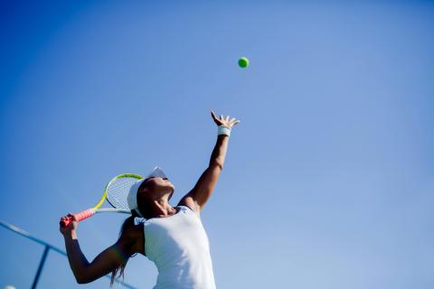 A woman is serving in a game of tennis. The camera looks up at her from the ground, and the sky is clear and blue in the background.