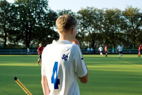 A teenage boy stands by the sidelines of a field hockey game with his back to the camera. All the action is far in the background.