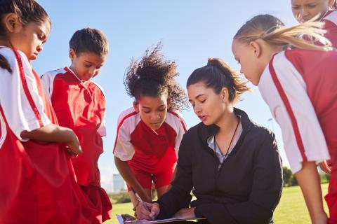 Junior football players surround their coach while she discusses what's on her clipboard.