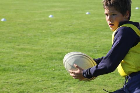 A boy holds a rugby ball during practice on a sunny, grassy playing field