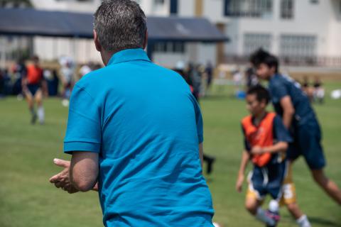 A parent on the sidelines claps for their child as they play football. The parent is wearing a blue polo shirt and the kids are blurred by the depth of field.