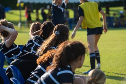 Girls on a rugby team sit on the bench while observing the field.