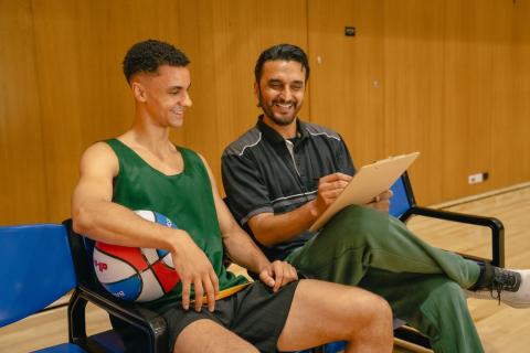 A coach and player sit on the sidelines of a basketball court smiling