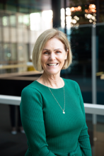A woman with fair skin and blonde hair smiles in a corpoate pose, wearing a green jumper and necklace.