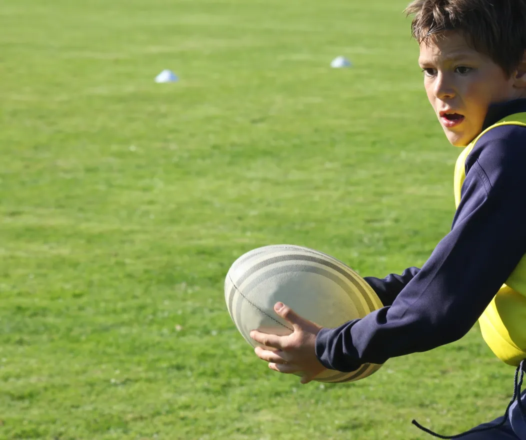 A boy holds a rugby ball during practice on a sunny, grassy playing field