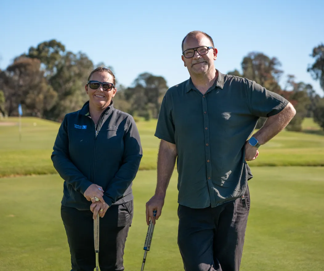 One women and one man with fair skin stand smiling on a golf course in the sun.
