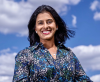 Dr Aish Ravi is a woman of South Asian descent. She is smiling and wearing a blue and white Liberty print business shirt. The background is a blue sky with white clouds.
