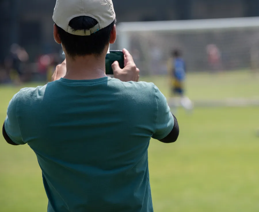 Man in white hat and green tshirt, facing away from camera, taking a photo of a kids soccer game