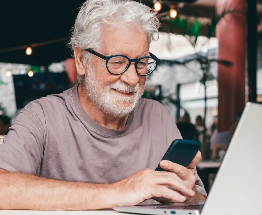 A man with silver hair and glasses smiles and works away on his laptop and smartphone