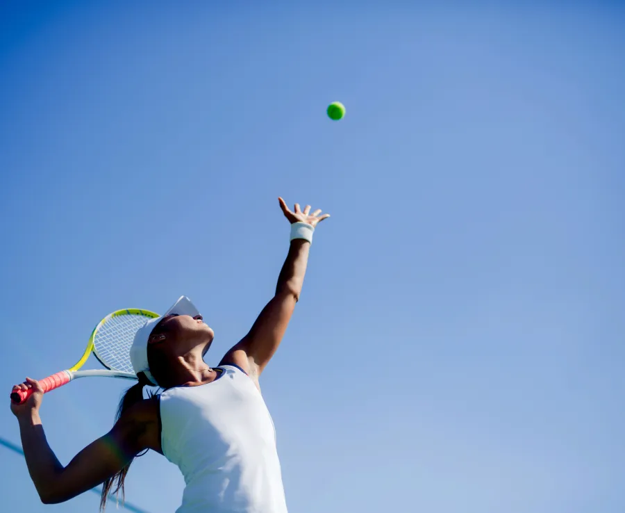 A woman is serving in a game of tennis. The camera looks up at her from the ground, and the sky is clear and blue in the background.