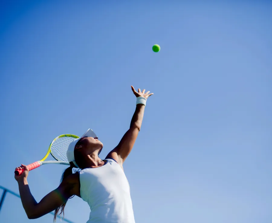 A woman is serving in a game of tennis. The camera looks up at her from the ground, and the sky is clear and blue in the background.
