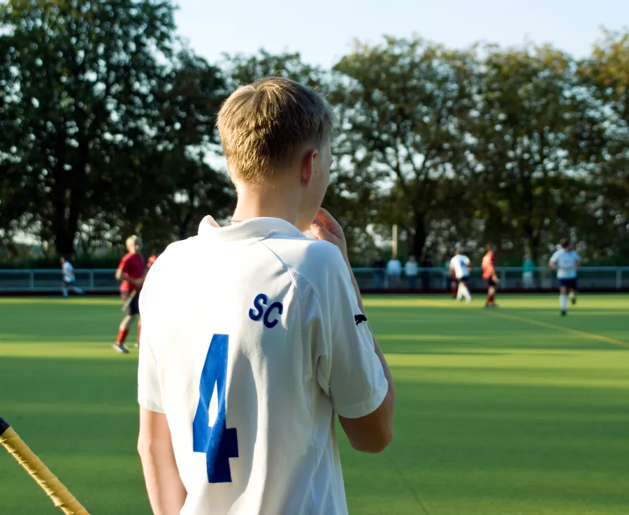 A teenage boy stands by the sidelines of a field hockey game with his back to the camera. All the action is far in the background.