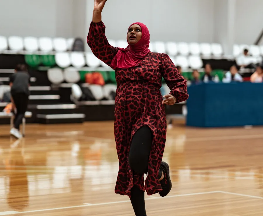Nabila is at the 3 point line, about to shoot the ball into the hoop. She is wearing a red hijab, a red and black midi dress, black leggings and black sneakers.