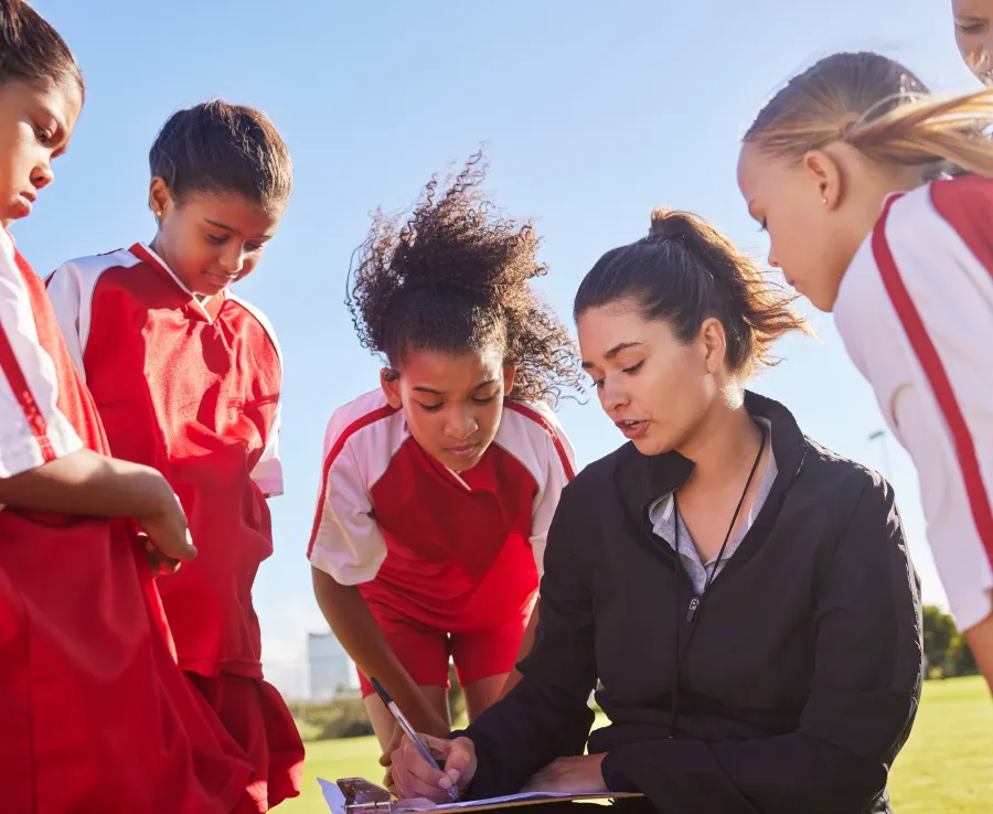 Junior football players surround their coach while she discusses what's on her clipboard.