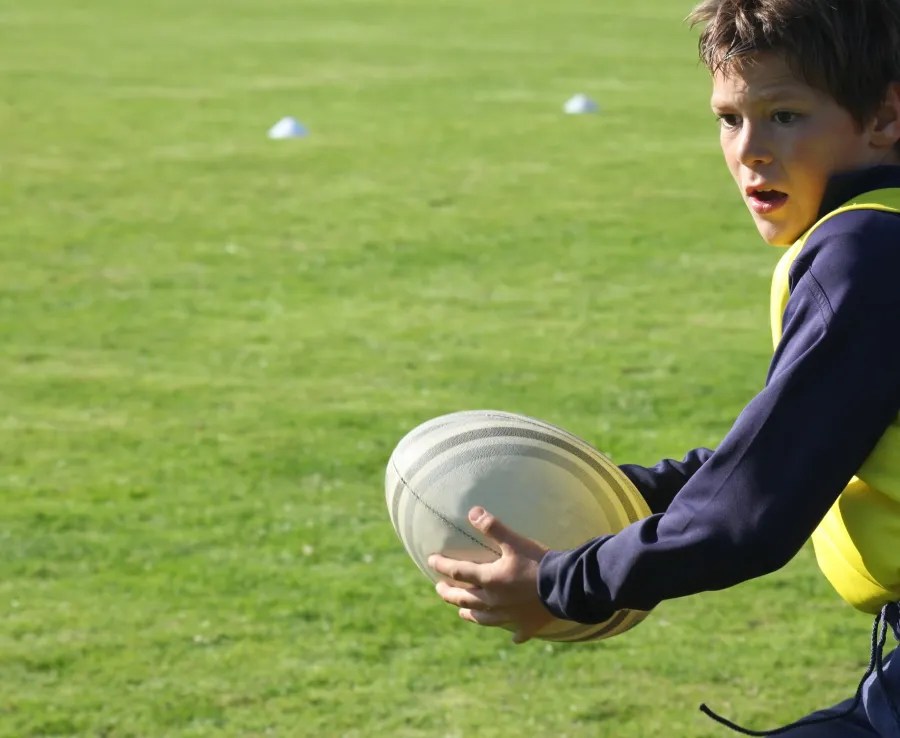 A boy holds a rugby ball during practice on a sunny, grassy playing field