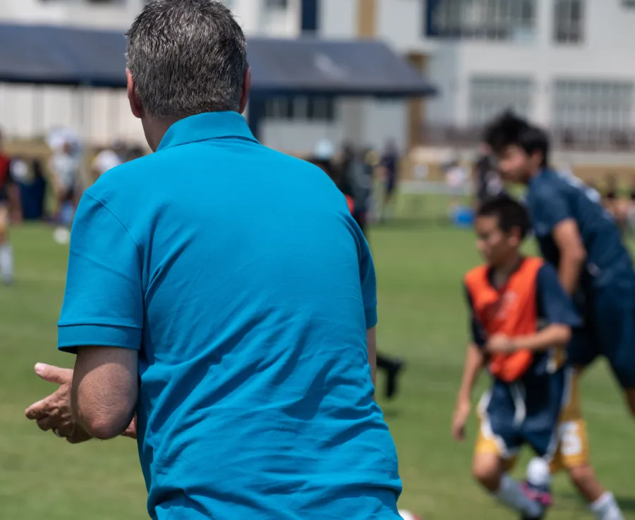 A parent on the sidelines claps for their child as they play football. The parent is wearing a blue polo shirt and the kids are blurred by the depth of field.
