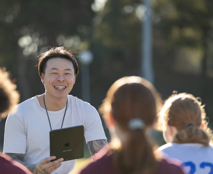 A male coach of East Asian appearance in a white tshirt holds an iPad and smiles while talking with female athletes who are listening attentively
