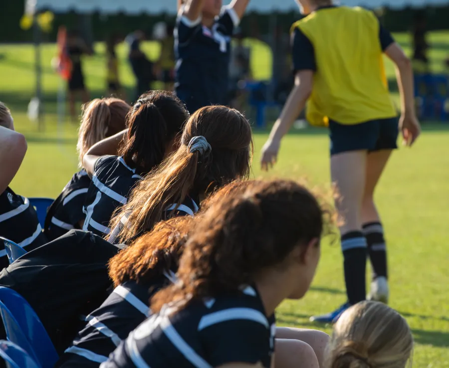 Girls on a rugby team sit on the bench while observing the field.