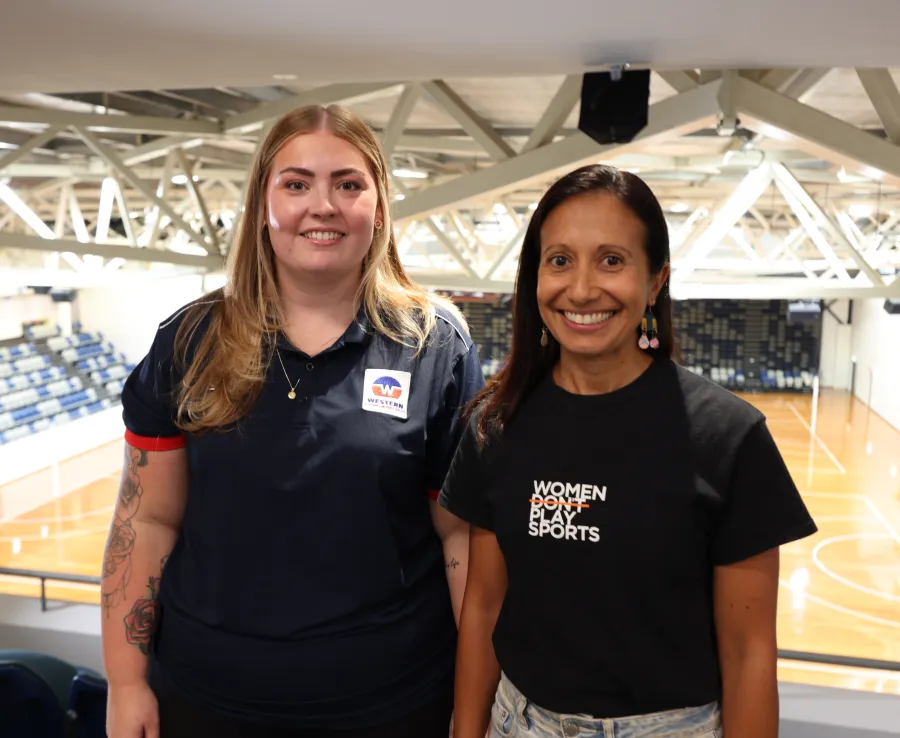 Two women smile at the camera inside an indoor stadium.