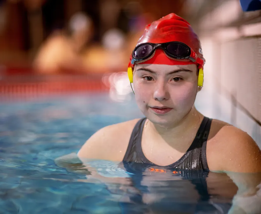 Young girl with Down's Syndrome wearing a swimming cap and goggles on her forhead in the water looks at the camera